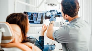 An orthodontists reviews an x-ray with a patient. Learn how DSOs and OSOs generate returns, where private equity fits in, and how doctor-owned models differ before you evaluate a partnership offer.